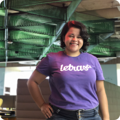 Jovem em pé com uma das mãos apoiadas na cintura. Ela está sorrindo e usa uma camiseta roxa com a logomarca do Letras. Sua pele é branca e seus cabelos pretos.