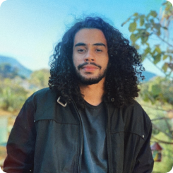 Jovem de pé esboçando um sorriso. Ao fundo, observa-se algumas árvores e montanhas. Ele usa uma jaqueta preta sobre uma camiseta também preta. Sua pele é negra e seus cabelos são longos e pretos.